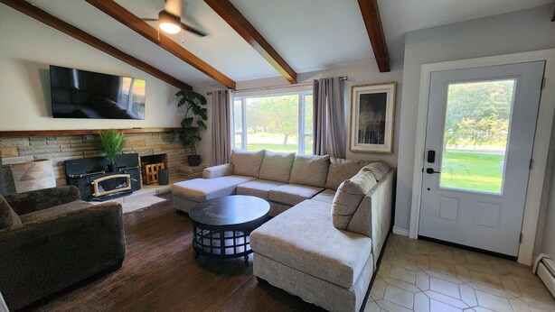 A living room with a sectional sofa, a coffee table, a fireplace, a flat-screen TV, and large windows offering a view of the outdoors. - Relax in Comfort! (Cornwall)