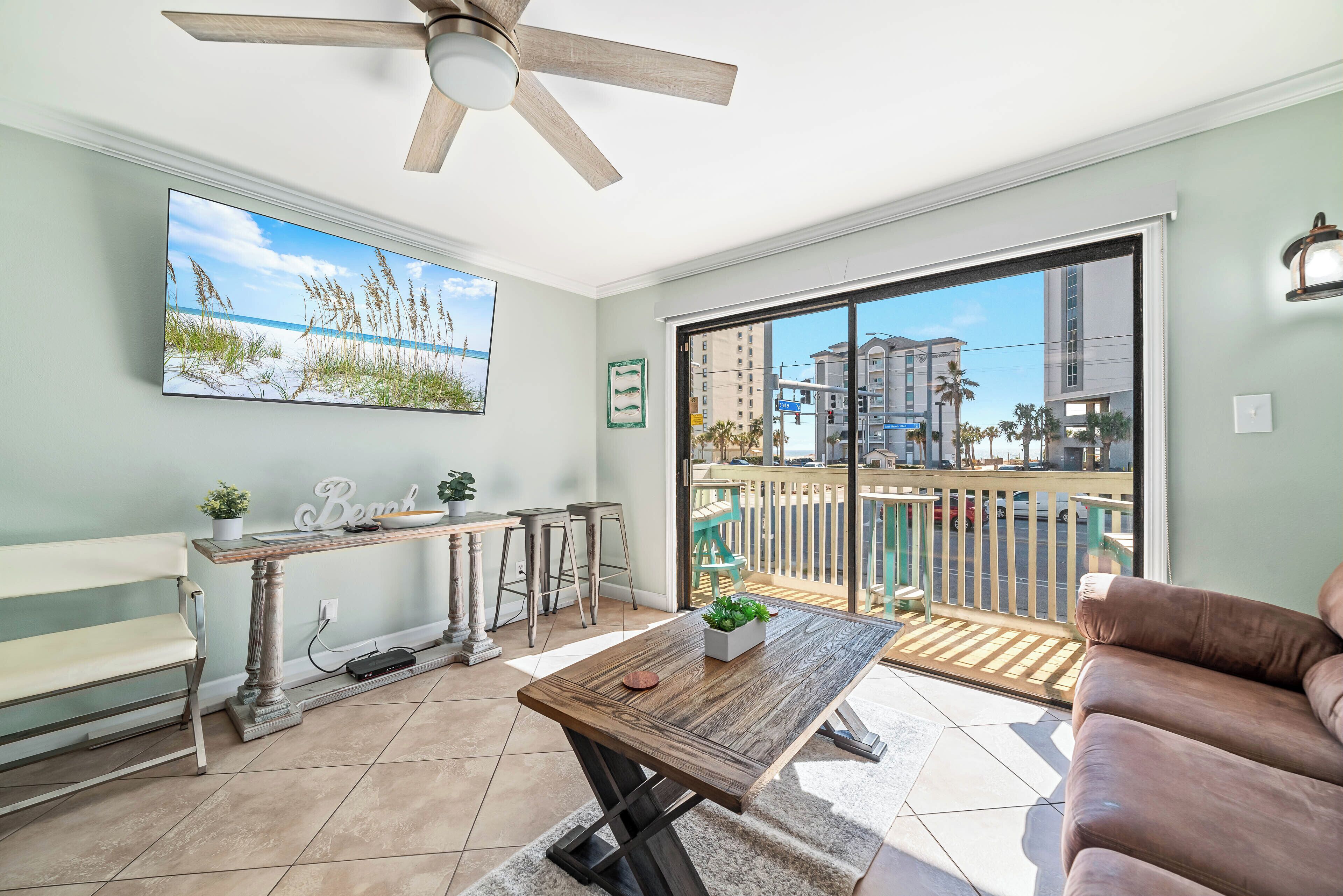 A living room with a wooden coffee table, a sofa, a bench, a ceiling fan, a large window, and a view of a building and palm trees outside.