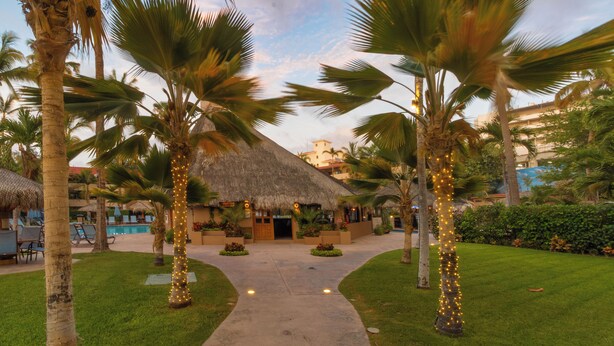 A resort with thatched-roof buildings, palm trees, and a pathway leading to a pool area.