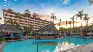 A resort pool area with a large building in the background, palm trees, and thatched umbrellas.