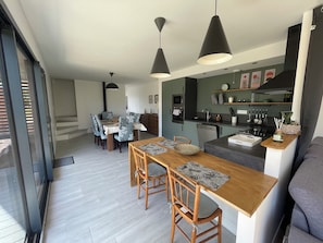 A modern kitchen with a dining area, featuring a wooden table, chairs, pendant lights, and a built-in oven. - Vacation home between land and sea (Coëtmieux)