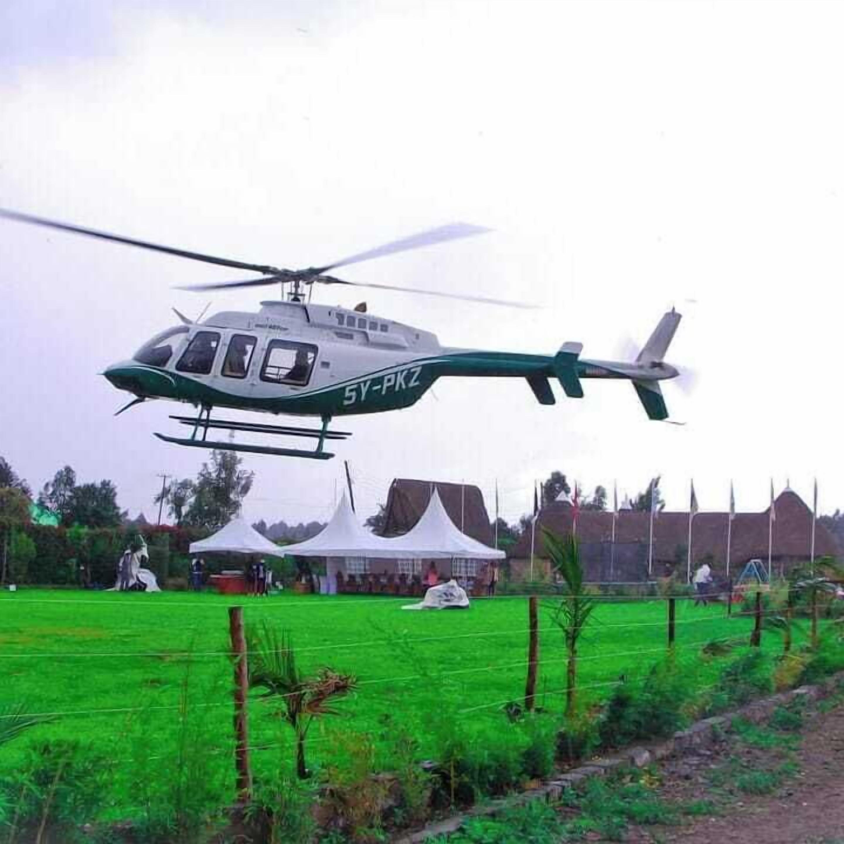 Een helikopter met registratienummer 5Y-PKZ vliegt boven een groen veld met tenten en mensen op de achtergrond.