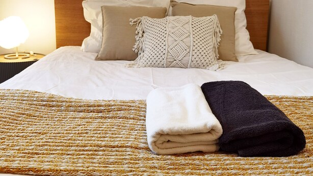 A neatly made bed with a white comforter, beige and white pillows, and a macrame throw. A folded white and dark blue towel is on the bed.