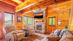 A cozy cabin interior with a stone fireplace, leather recliners, and a large window.