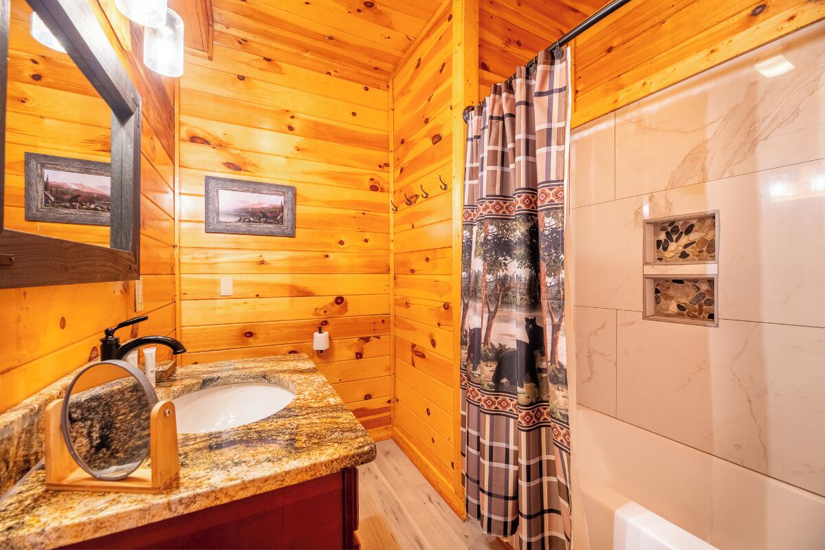 Bany d'una cabana de fusta amb un lavabo de pedra granat, un mirall, una cortina de bany amb una escena de bosc i una petita estant amb pedres decoratives.
