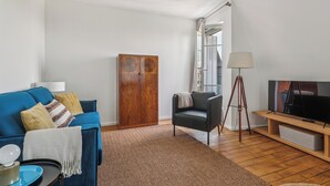 A living room with a blue sofa, a wooden wardrobe, a black armchair, a TV on a wooden stand, and a lamp. - Charming 1BR in Le Marais - Bretagne (Paris)