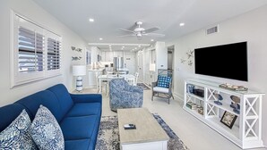 Una sala de estar moderna con un sofá azul, una mesa de centro, un televisor sobre un mueble blanco y un área de comedor al fondo.