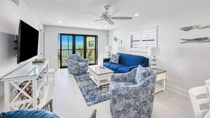 Una sala de estar moderna con una televisión de pantalla plana, ventilador de techo, sofá azul, sillones estampados y un ventanal con vista al océano.
