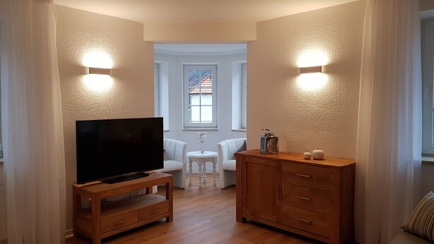 Una sala de estar con un televisor sobre un mueble de madera, un sofá blanco, una mesita redonda con un florero de vidrio y un armario de madera con cajones.