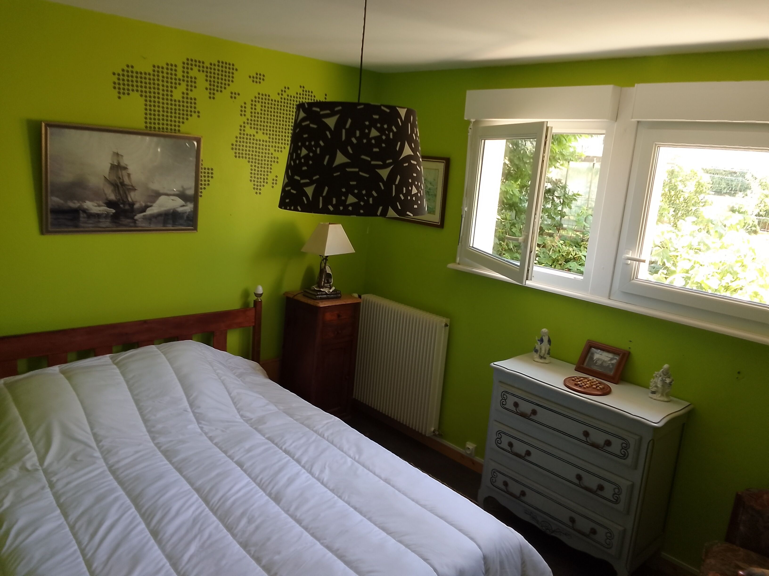 Une chambre à coucher avec un lit, une commode, une carte du monde accrochée au mur, une lampe et une fenêtre donnant sur un paysage verdoyant.