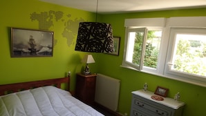 Une chambre à coucher avec un lit, une commode, une carte du monde accrochée au mur, une lampe et une fenêtre donnant sur un paysage verdoyant.