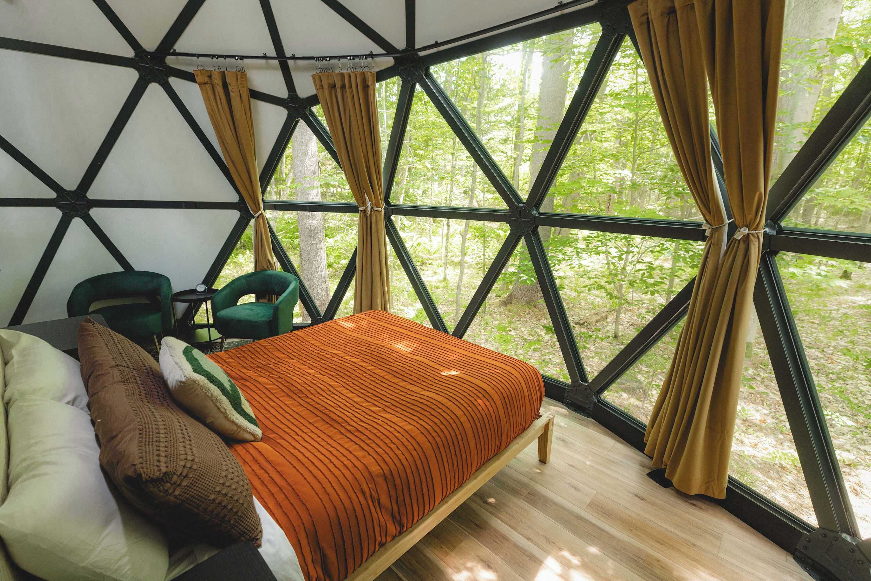 A room with a bed, a chair, and a view of a forest through large windows.