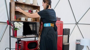 Une personne est en train de disposer des objets sur une étagère dans une pièce dont le mur est blanc et présente un motif géométrique, le sol est en bois et un appareil électroménager est de couleur rouge.