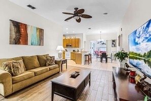 A modern living room with a sofa, coffee table, and TV. There is a kitchen in the background with cabinets and a dining area. - Pool Table | Conservation View | Near Disney 354 (Davenport)