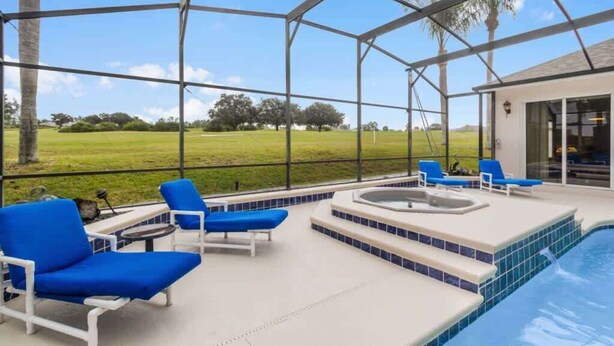 Zona de piscina exterior techada con sillas reclinables azules, un jacuzzi y vistas a un campo verde.