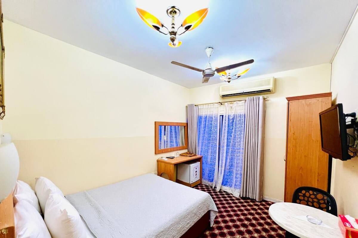 A hotel room with a bed, a desk, a chair, a table, a ceiling fan, and an air conditioning unit.