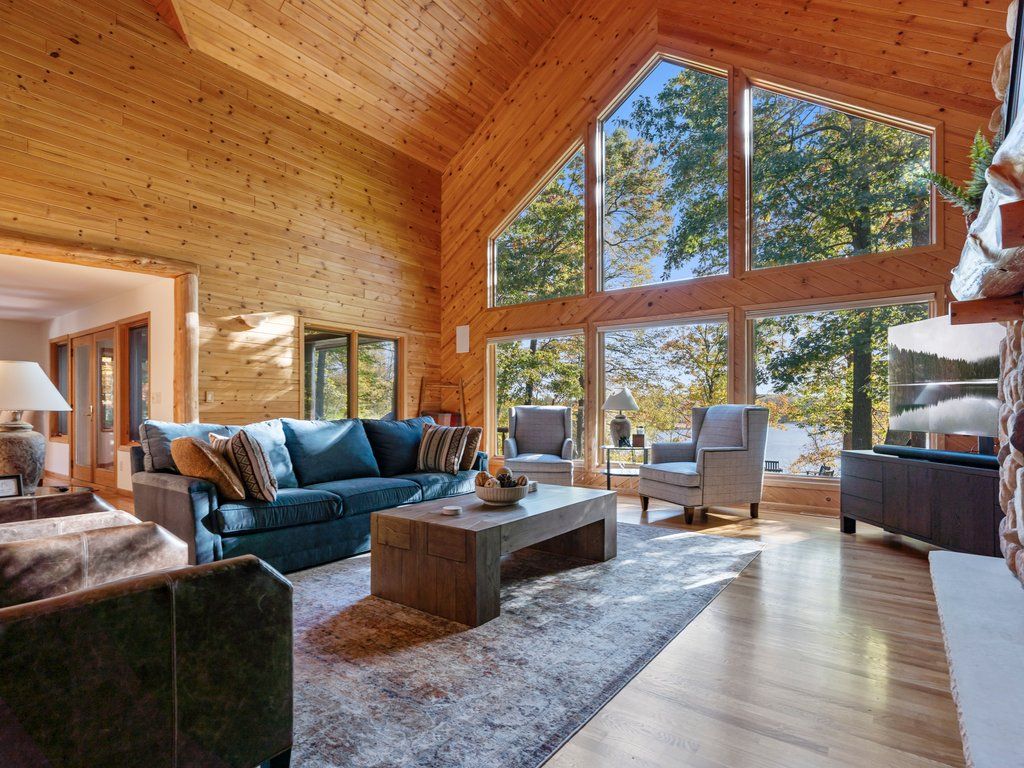 A spacious living room with a large wooden ceiling, a blue sofa, a coffee table, and a view of trees through large windows.