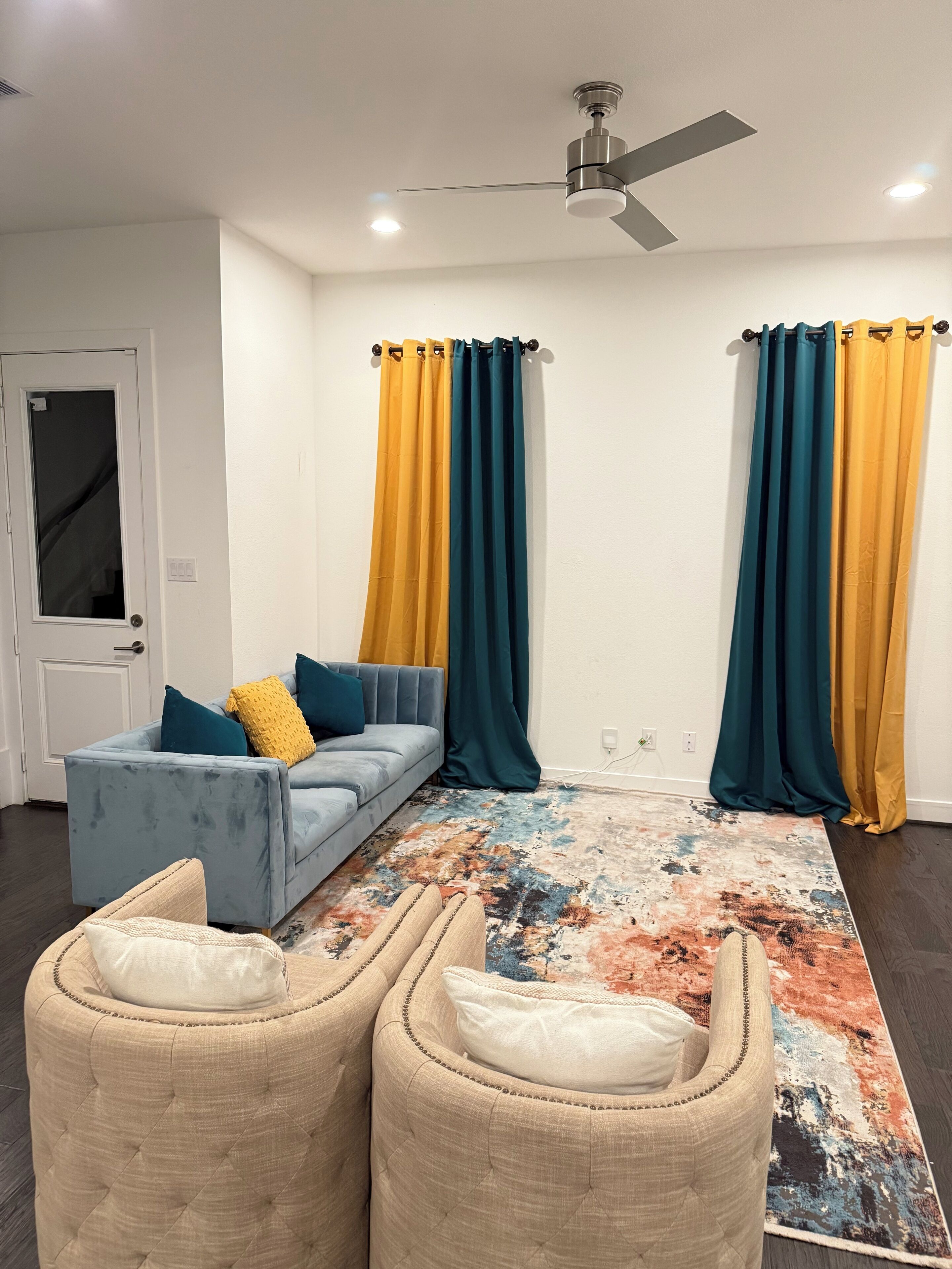 Een moderne woonkamer met een grijze bank, twee fauteuils, een rug met een patroon en teal- en geelkleurige gordijnen.