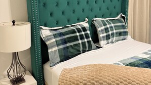 Ein Bett mit einem grünen, bezogenen Kopfteil, zwei karierten Kissen, einer beige farbenen Tagesdecke und einem weißen Nachttisch mit einer Lampe.