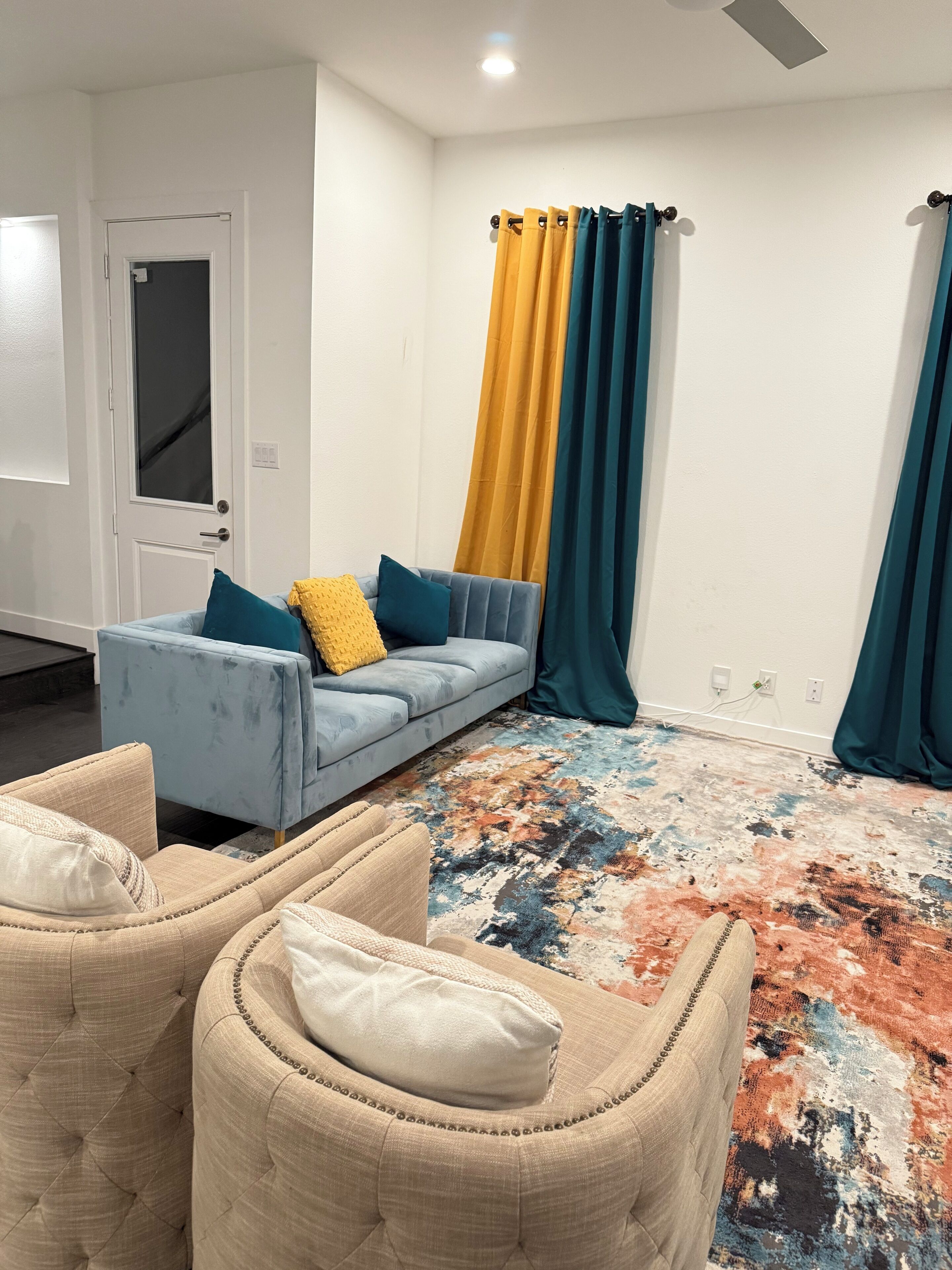 A living room with a light blue sofa, beige armchairs, a colorful rug, and teal curtains.