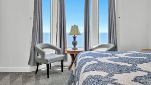 Habitación de hotel con un amplio ventanal, dos sillas, una mesita con una lámpara y una cama con colcha estampada.