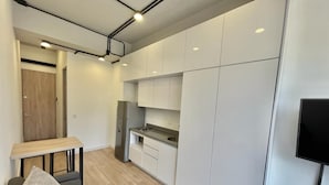 Cocina compacta con alacenas blancas, un refrigerador empotrado y un pequeño comedor con mesa y sofá.