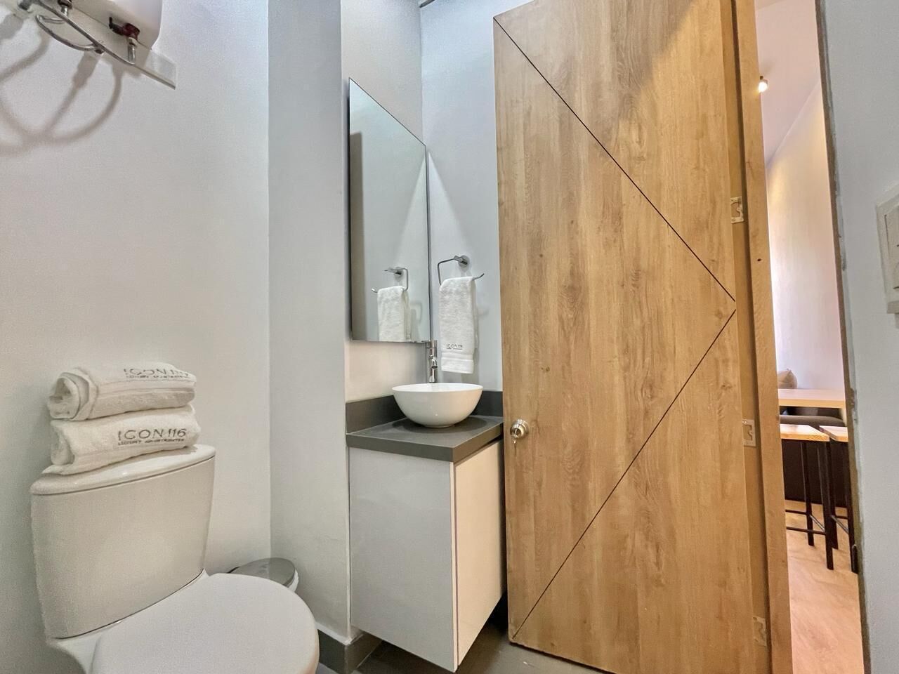 Un baño moderno con inodoro, lavamanos y espejo. Hay toallas apiladas sobre el inodoro. Se ve una puerta de madera al fondo.