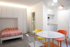 A compact room with a bed, a round table with chairs, a kitchenette, and a door leading to another room.