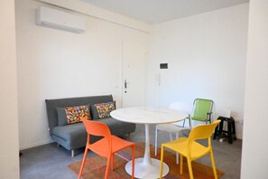 Une petite pièce avec un canapé gris, des coussins colorés, une table blanche et deux chaises.