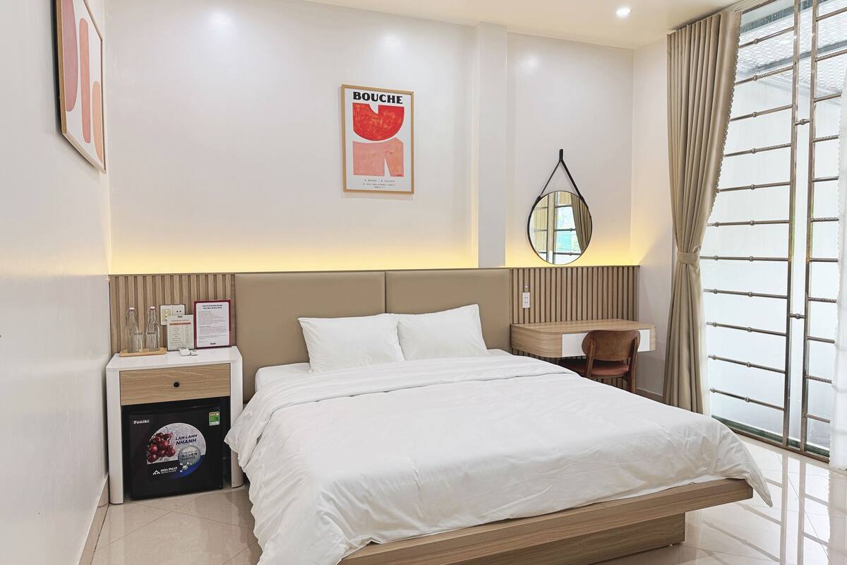 A hotel room with a bed, a desk, a chair, a minibar, and a large window with curtains.