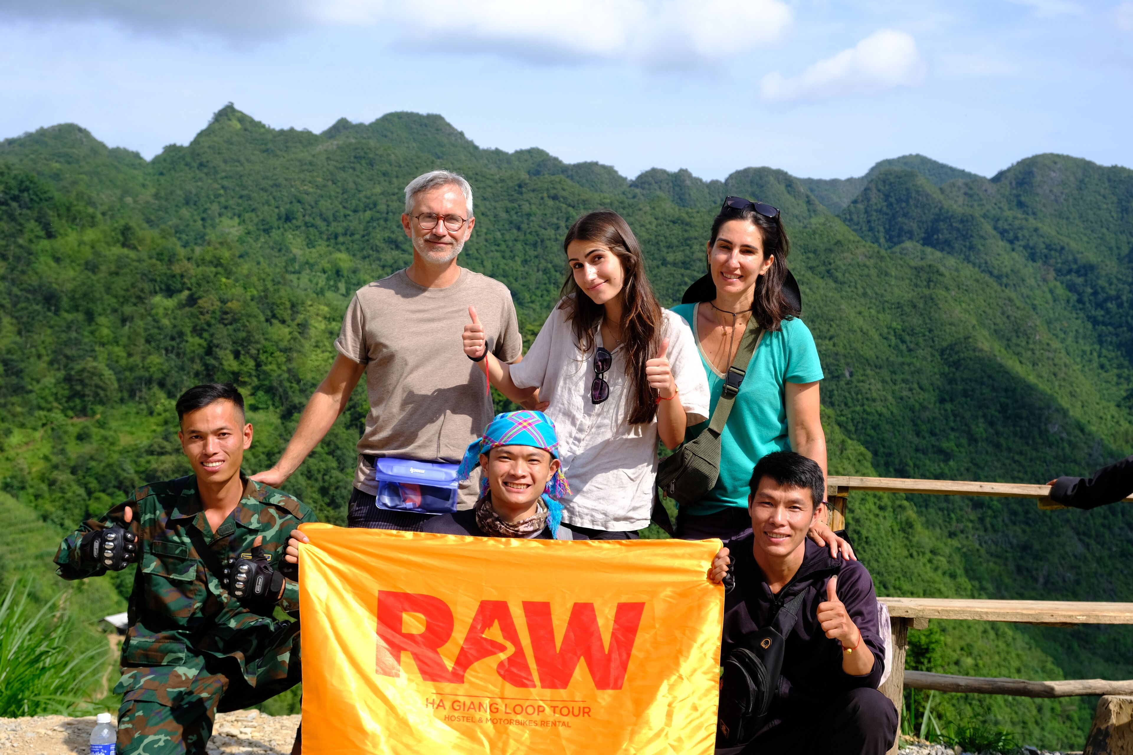 A group of people standing on a wooden platform with a banner that reads 'RAW HA GIANG LOOP TOUR HOSTEL & MOTORBIKE RENTAL'.