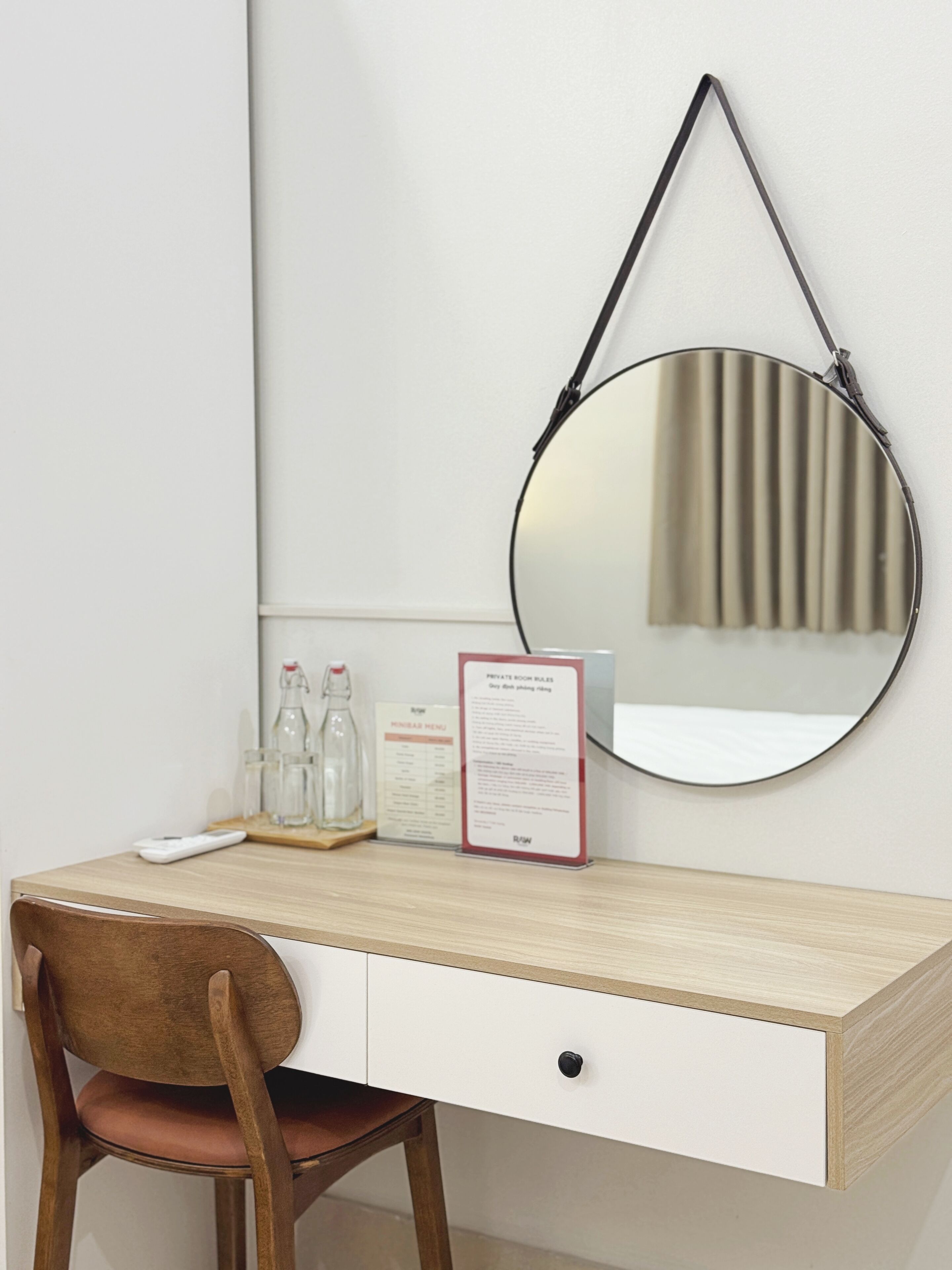 A wooden desk with a white drawer, a round mirror, a bottle of water, a glass, and a menu sign.