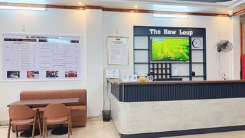 Un mostrador de recepción con un cartel que dice 'The Raw Loop', un tablón de anuncios con horarios de autobús y una zona de estar con un sofá y sillones marrones.