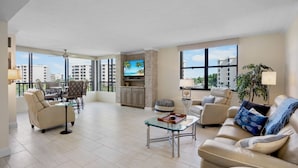 Amplia sala de estar con sofá, sillones, una mesa de centro y televisor. Posee un balcón con vistas a edificios y palmeras.