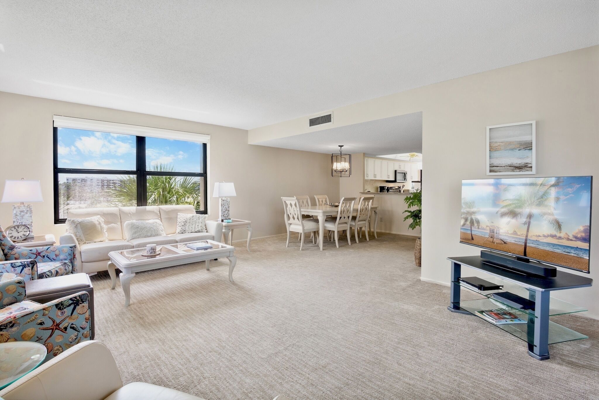 A spacious living room with a large window, a dining area, and a kitchen in the background.