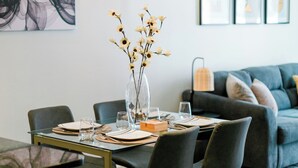 Un comedor con una mesa de vidrio, cuatro sillas y un jarrón con flores. Se ve un sofá y una lámpara al fondo.