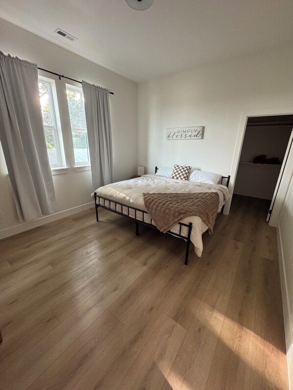 Une chambre à coucher avec un lit, un plancher en bois, une fenêtre avec des rideaux, un panneau fixé au mur et un placard.