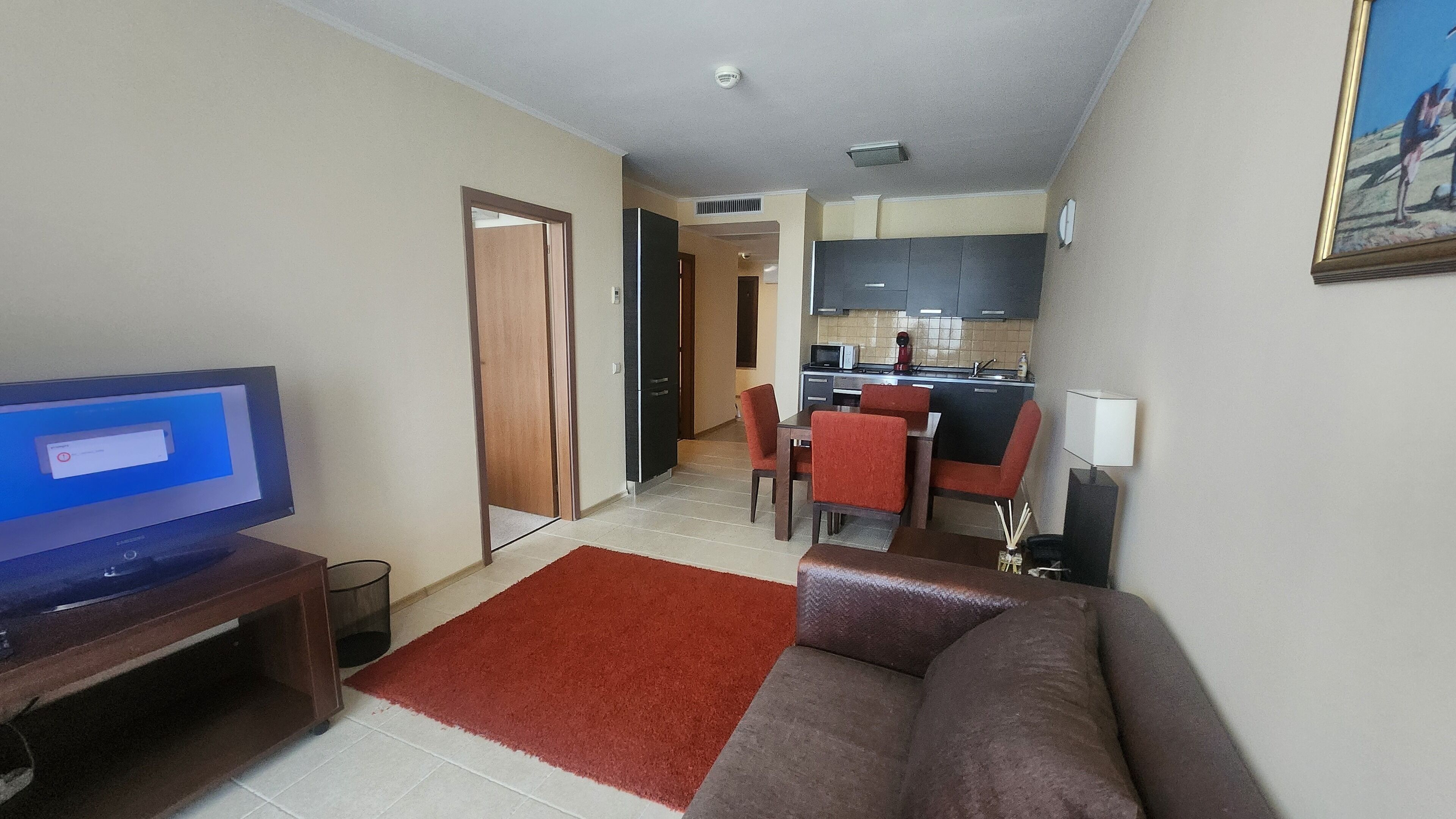 Una sala de estar con televisor, un tapete rojo y un sofá. Se ve una cocina al fondo con armarios y un microondas.