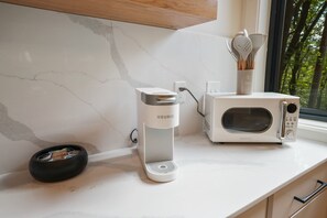 A kitchen with a white countertop, a Keurig coffee maker, a microwave, and a window with a view of trees.