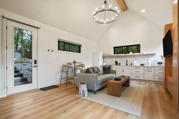 A modern living room with a grey sofa, wooden coffee table, and a chandelier. There is a kitchen area with white cabinets and a TV mounted on the wall.