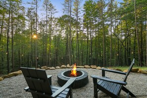 A circular fire pit surrounded by wooden chairs in a forest setting.