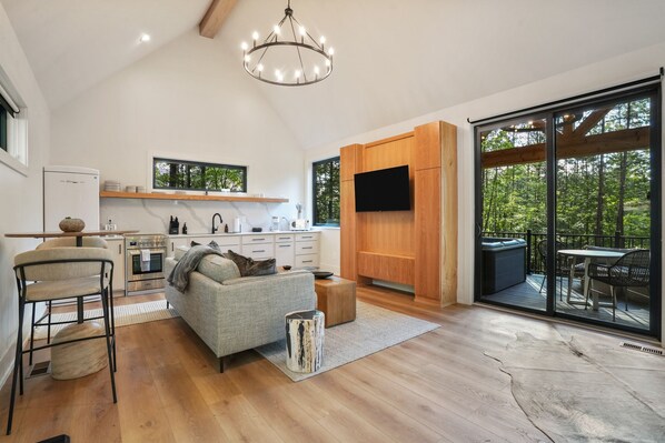 A spacious living area with a kitchen, a grey sofa, a wooden dining table, and a large sliding glass door leading to an outdoor deck.