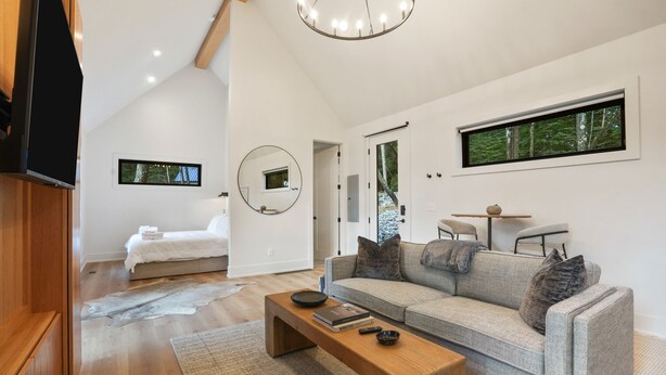 A modern living room with a sofa, coffee table, and a TV. There is a bedroom with a bed and a mirror. The room has large windows and a wooden ceiling. - Nature's Loft | Hocking Hills, New Modern Cabin (Rockbridge)
