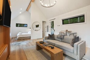 A modern living room with a sofa, coffee table, and a TV. There is a bedroom with a bed and a mirror. The room has large windows and a wooden ceiling.
