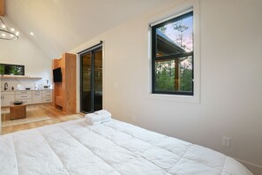 Une chambre avec un grand lit, une armoire en bois, un coin cuisine et une fenêtre donnant sur les arbres.