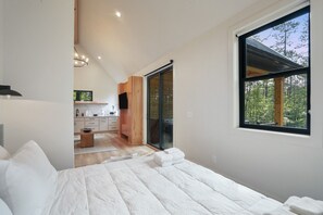 A bedroom with a large bed, a view of a kitchen, and a sliding glass door leading to an outdoor area.