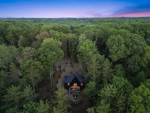 Une vue aérienne d’une cabane entourée d’une forêt dense, avec un ciel dégagé et des collines lointaines.