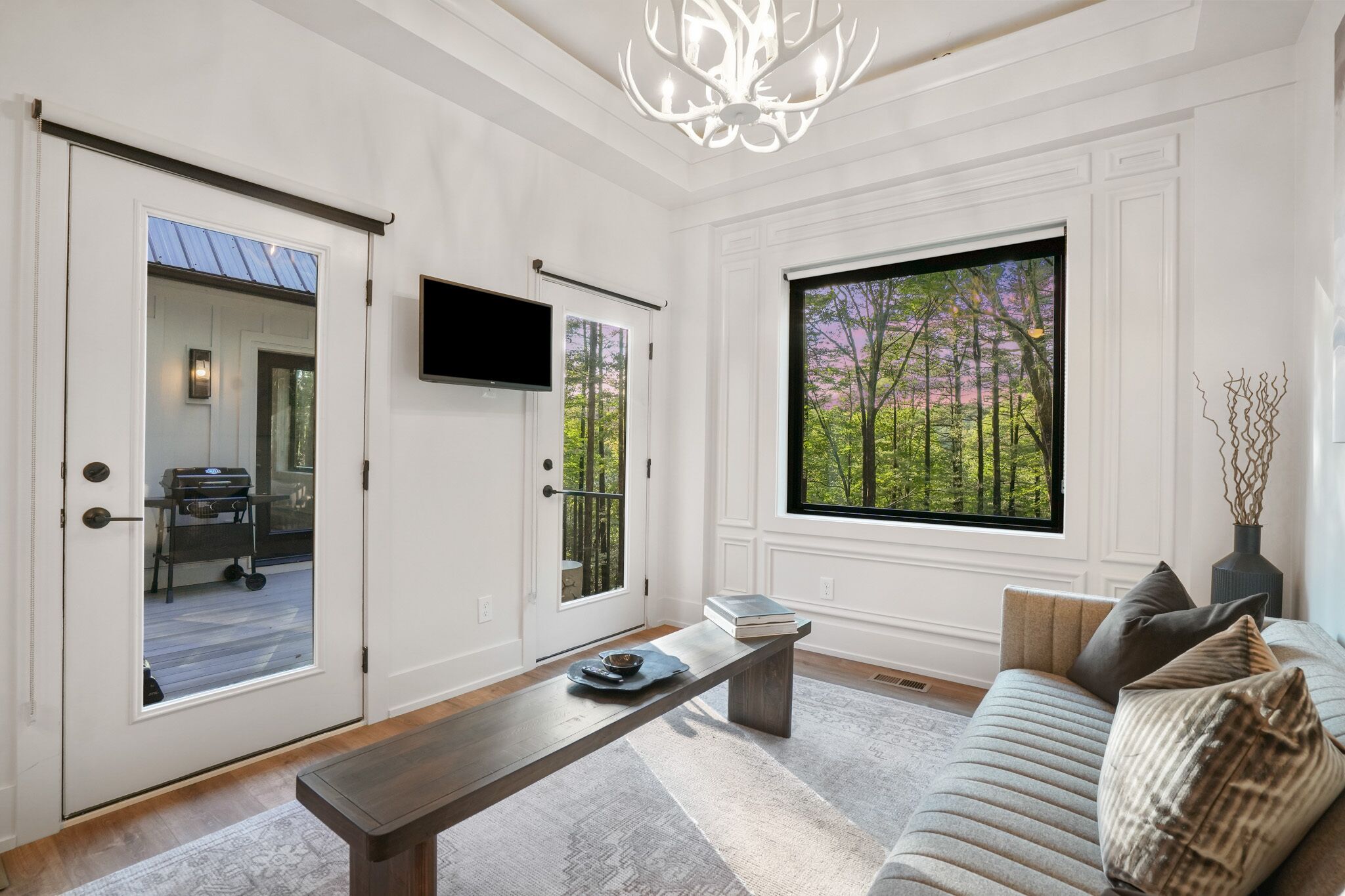 Un salón moderno con un sofá, una mesa de centro y un banco. Hay un televisor montado en la pared y un ventanal con vistas a árboles.