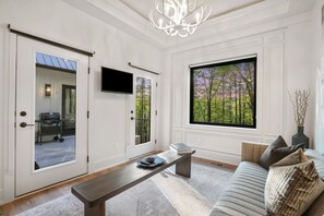 Un salón moderno con un sofá, una mesa de centro y un banco. Hay un televisor montado en la pared y un ventanal con vistas a árboles.