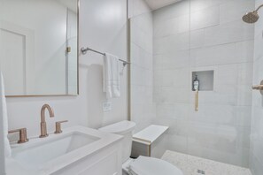 Un baño moderno con un lavamanos blanco, dos llaves, un espejo, un perchero para toallas, una ducha con cabezal de ducha manual y un estante pequeño con artículos de aseo.